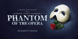 The Phantom, in a dark cape and mask, stands in a dimly lit theatre, surrounded by ornate decor.