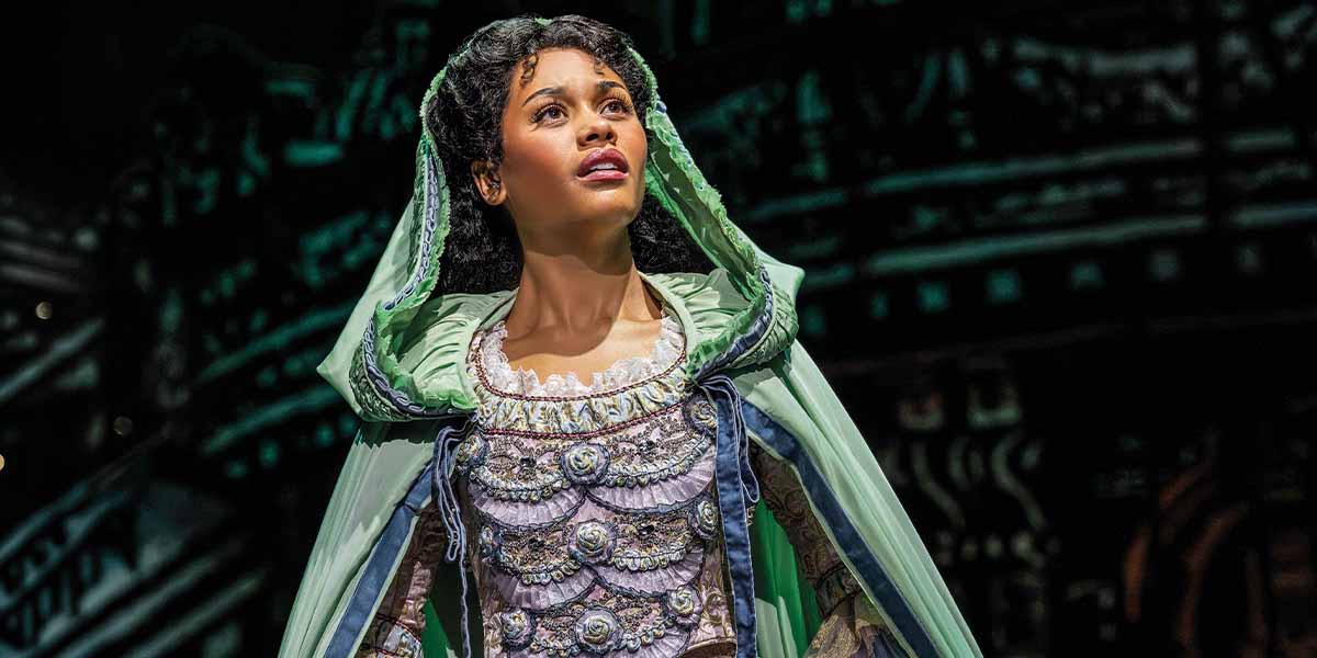 Beatrice Penny-Touré as Christine, wearing an ornate green costume, gazes upwards against a dark background.