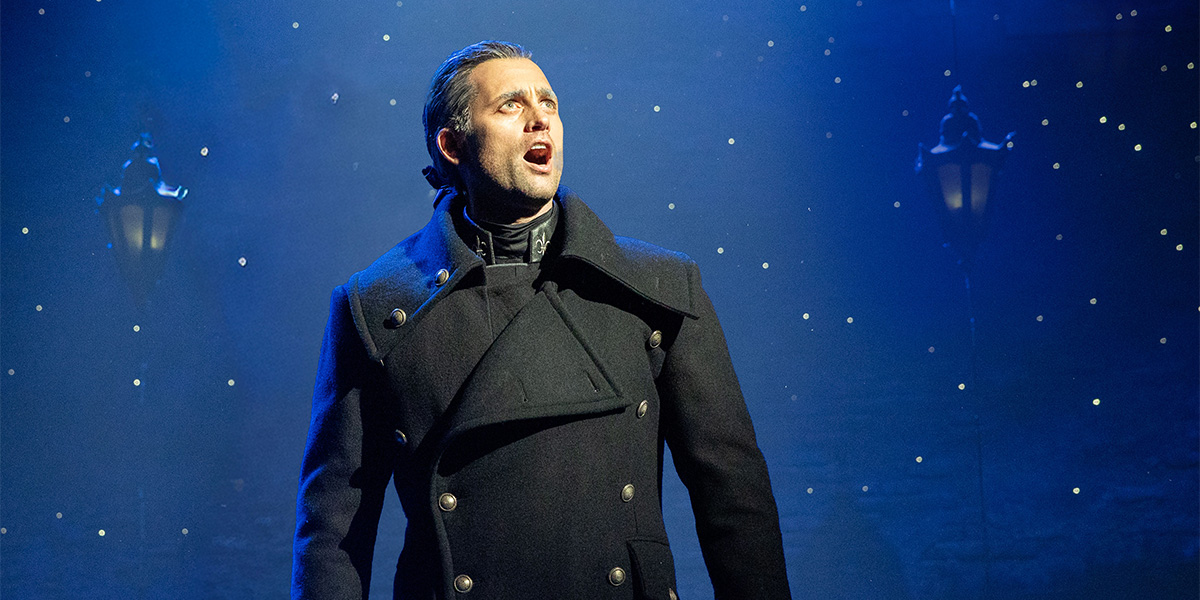 A man in a dark coat sings passionately under blue lighting, with twinkling stars in the background.