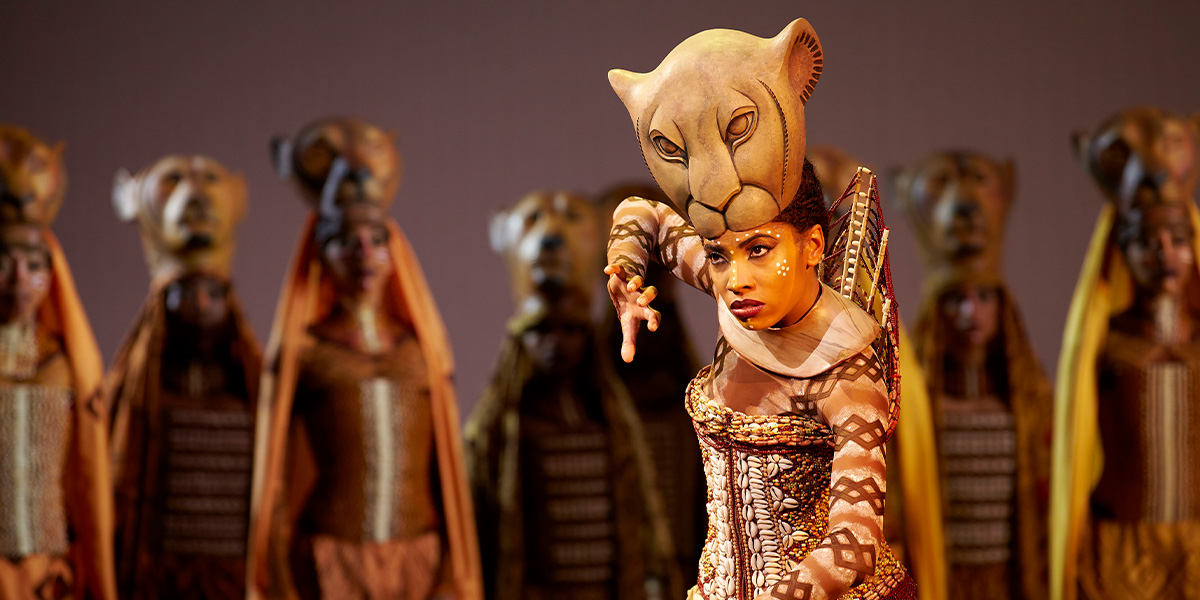 Daniela Pobega as Nala, wearing a brown costume, stands gracefully on stage with a dramatic backdrop.