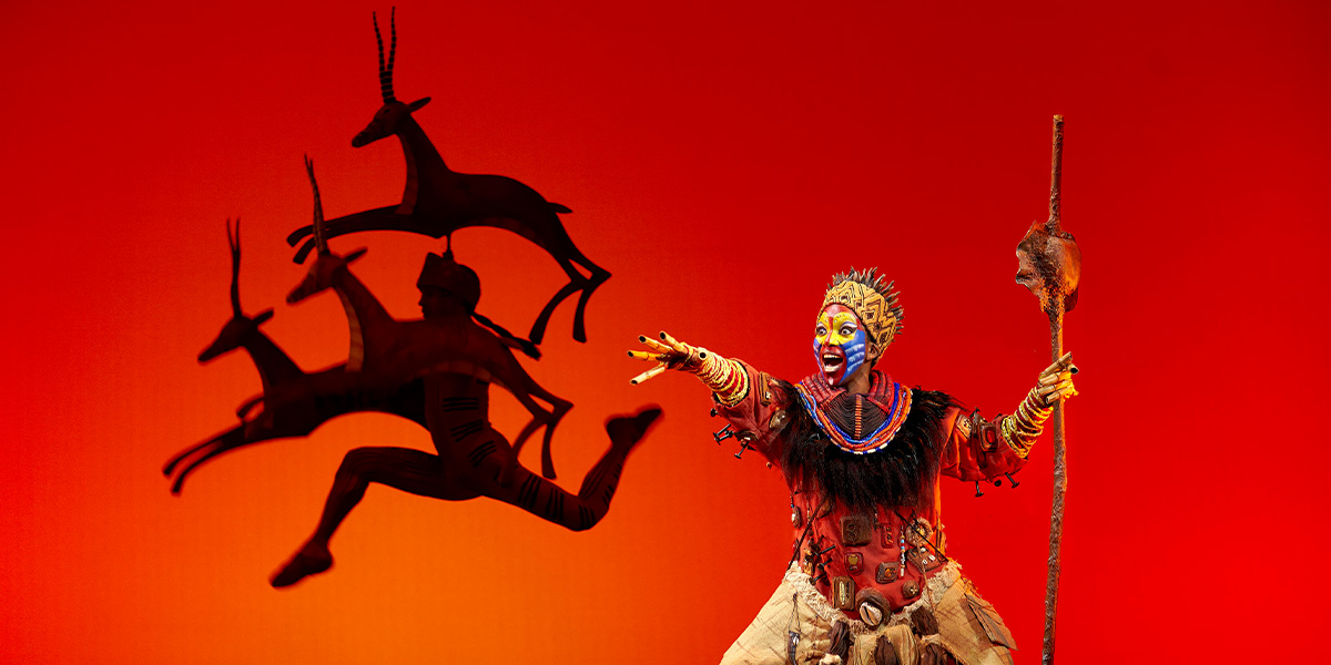 Rafiki, adorned in vibrant costume, gestures dramatically against a bright orange background with gazelle shadows.