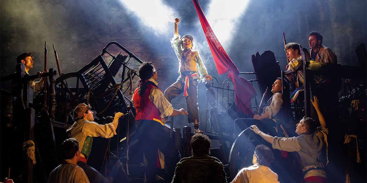 A group of performers in period costumes, with one raising a fist, amidst dramatic lighting and props.