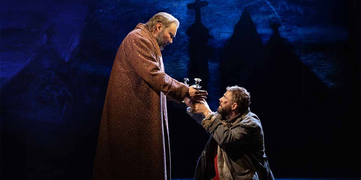 Two actors in period costumes interact on stage, one handing a silver object to the other.