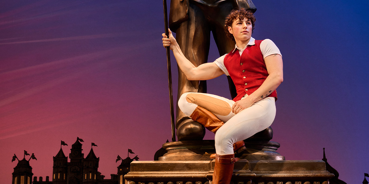 Fiyero, dressed in a green jacket, poses confidently with a playful expression against a dark background.