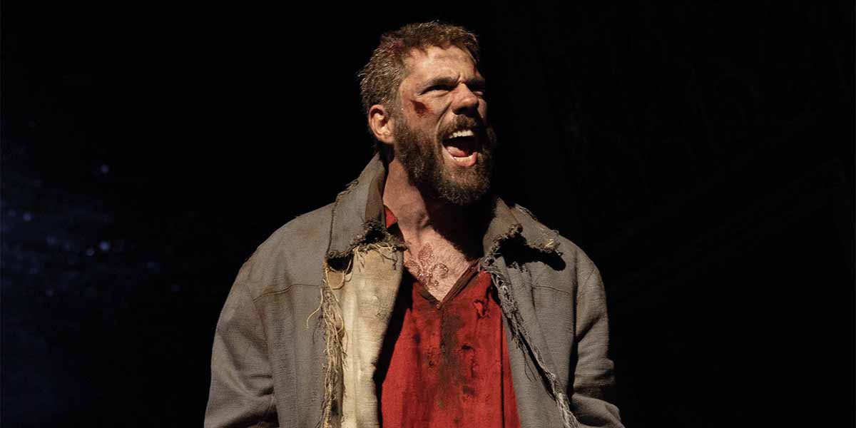 Ian McIntosh as Valjean, shouting passionately, wearing a tattered grey coat over a blood-stained red shirt.