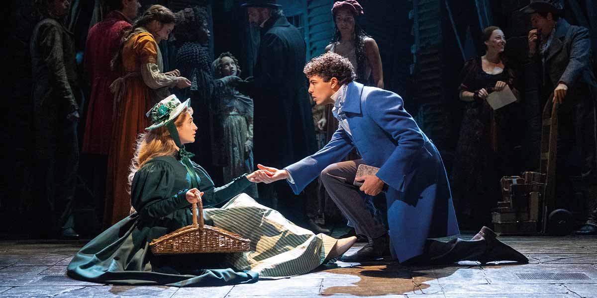 Izzi Levine as Cosette sits on the floor, while Thiago Phillip Felizardo as Marius kneels, holding hands.