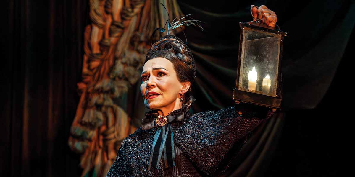 Joanna Riding as Madame Giry, holding a lantern, wearing a dark, textured costume, with dramatic backdrop.
