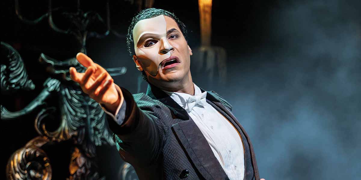Jordan Donica as the Phantom, wearing a half mask, gestures dramatically against a smoky, dark background.
