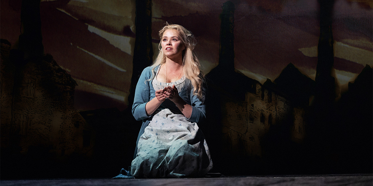 A woman kneels on stage, wearing a light dress and blue cardigan, with a dramatic backdrop.