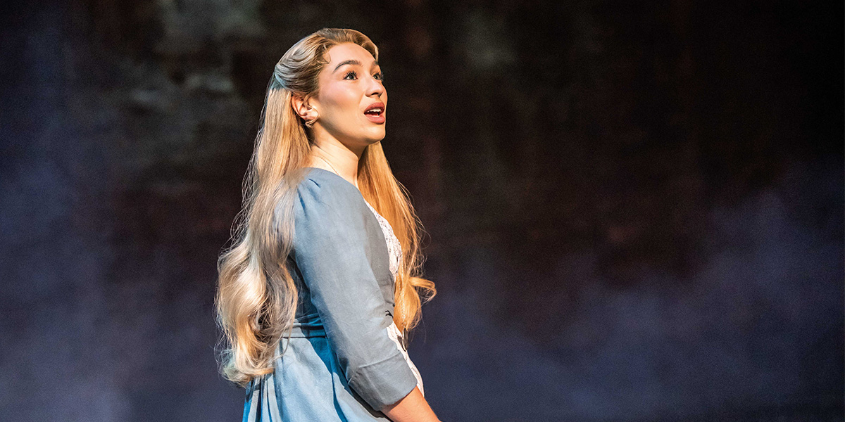 Martha Kirby as Fantine, wearing a tattered dress, with a distressed expression against a dark background.