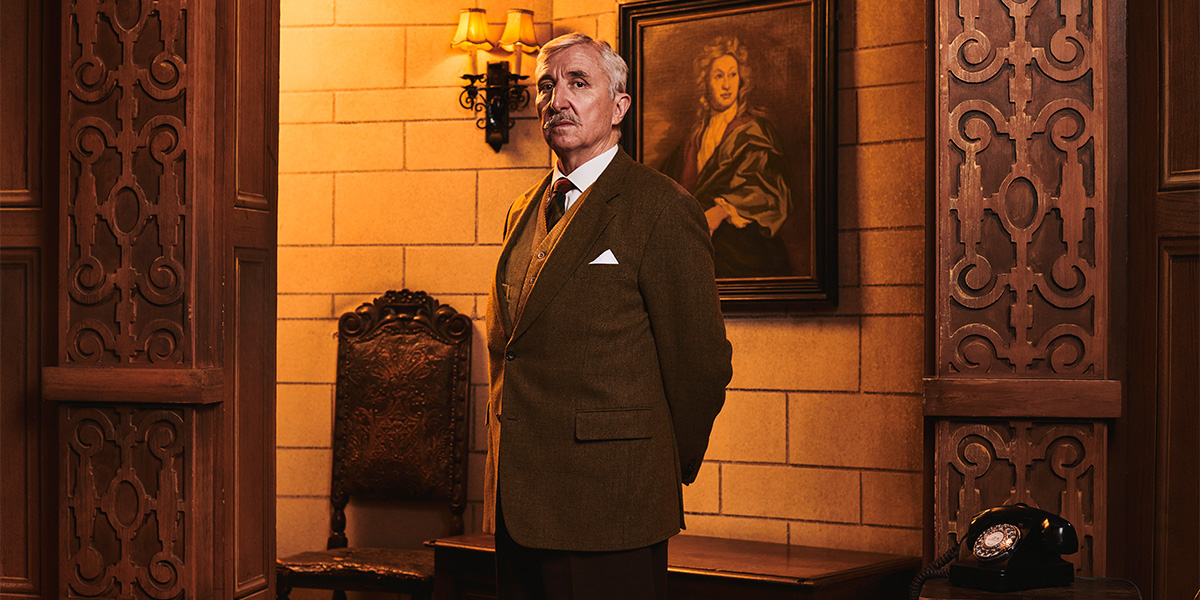 A scene from The Mousetrap featuring actors in period costumes on stage, with dramatic lighting and a wooden set.