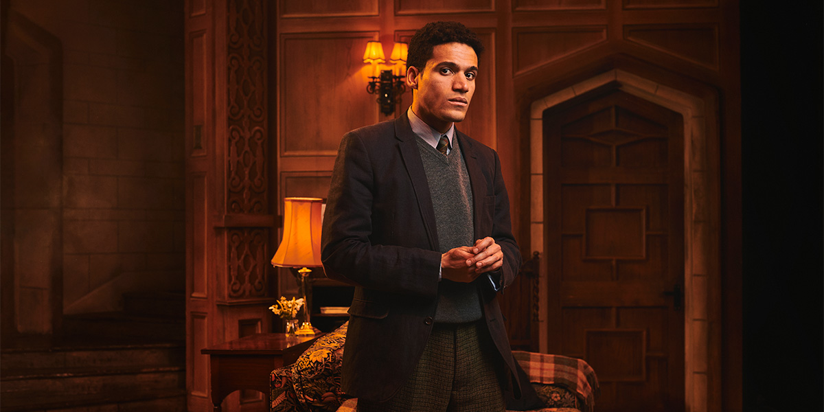 A man in formal attire stands in a warmly lit room, with wooden walls and a lamp beside him.