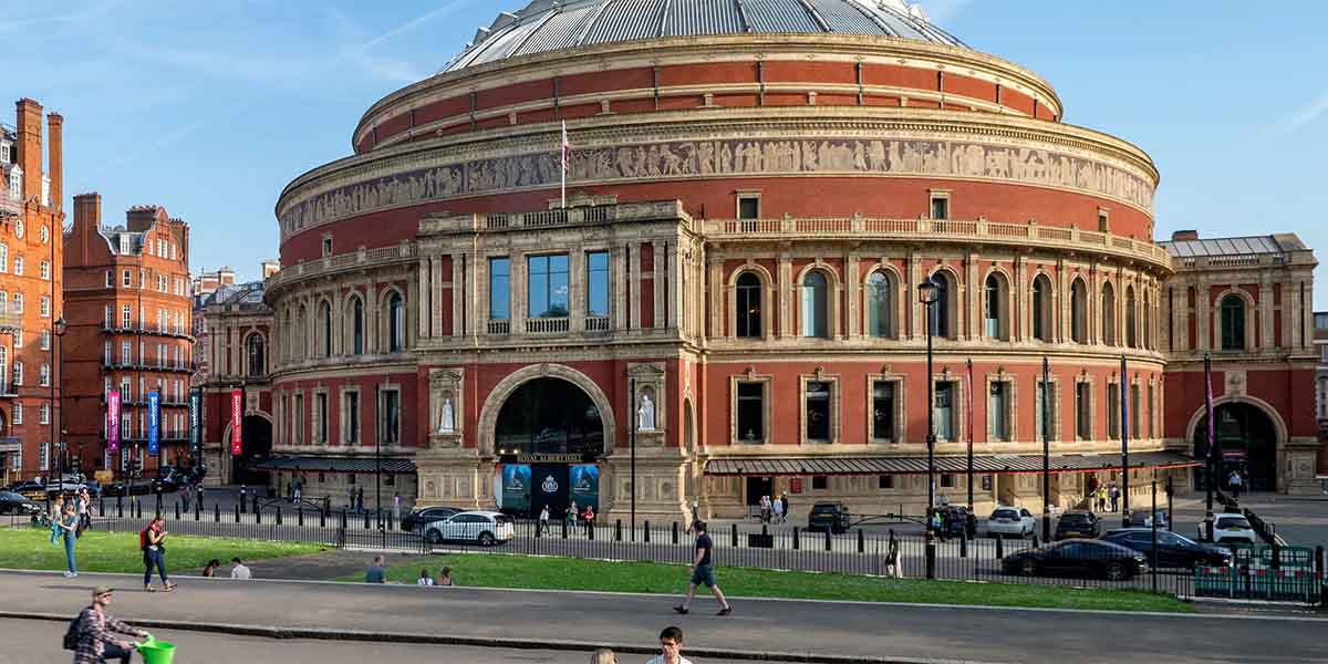 The Royal Albert Hall exterior, featuring its iconic circular dome and ornate brickwork, under a clear blue sky.