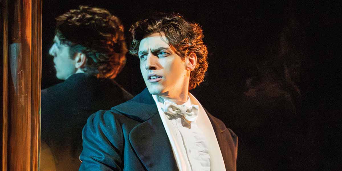Rhys Whitfield as Raoul, wearing a black suit and white bow tie, gazes thoughtfully with a reflective background.