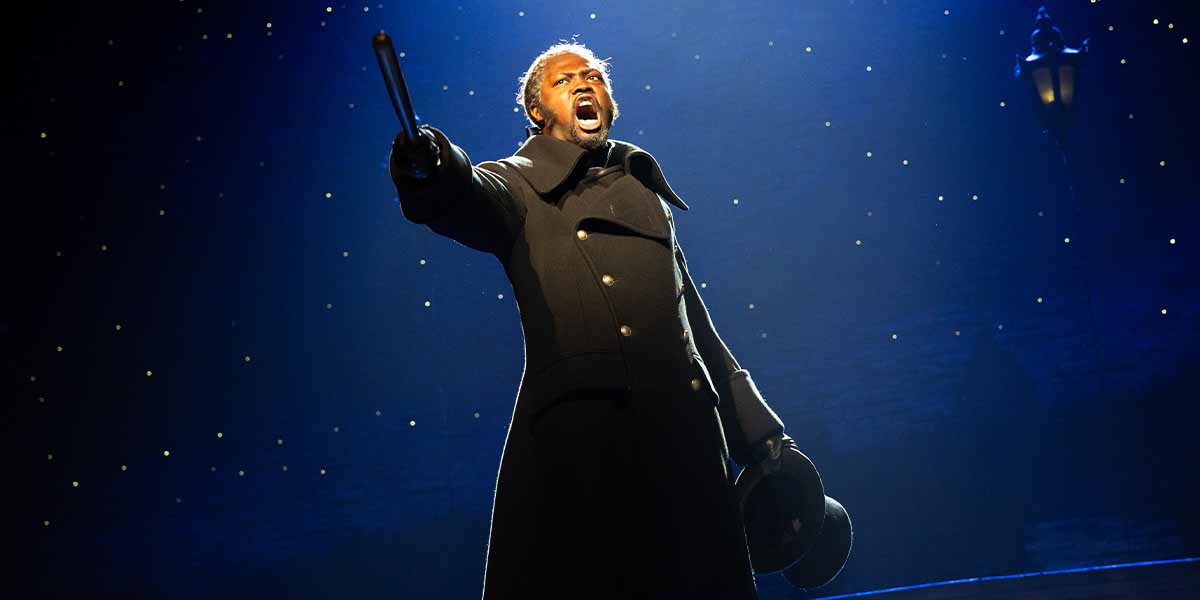Sam Oladeinde as Javert passionately sings, holding a baton, against a starry blue background.