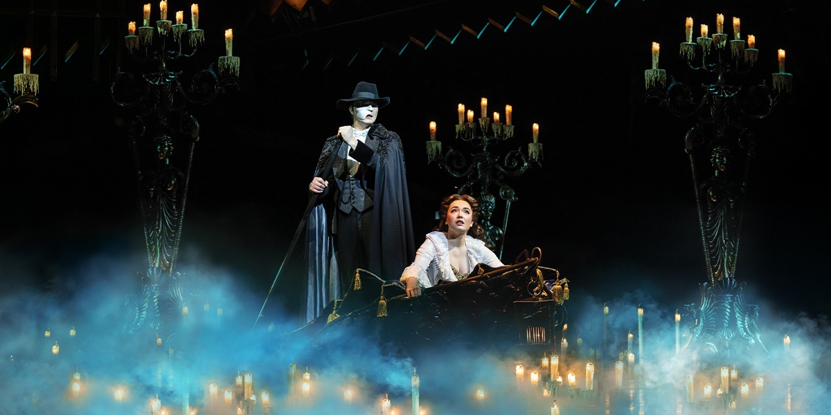 The Phantom stands behind Christine, who sits in a boat surrounded by candles and fog.