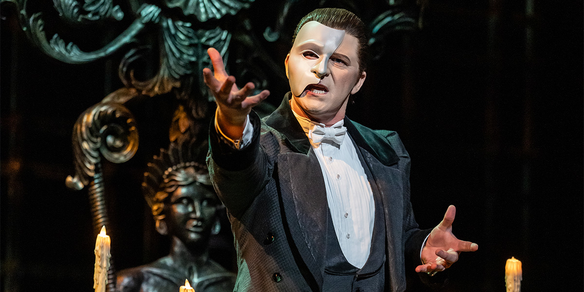 A man in formal attire and a half-mask gestures dramatically, surrounded by candles and ornate decor.