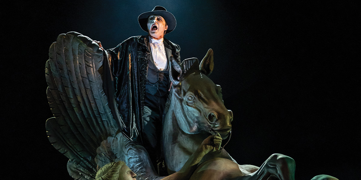 A man in a black cape and hat dramatically poses atop a large, winged horse sculpture.