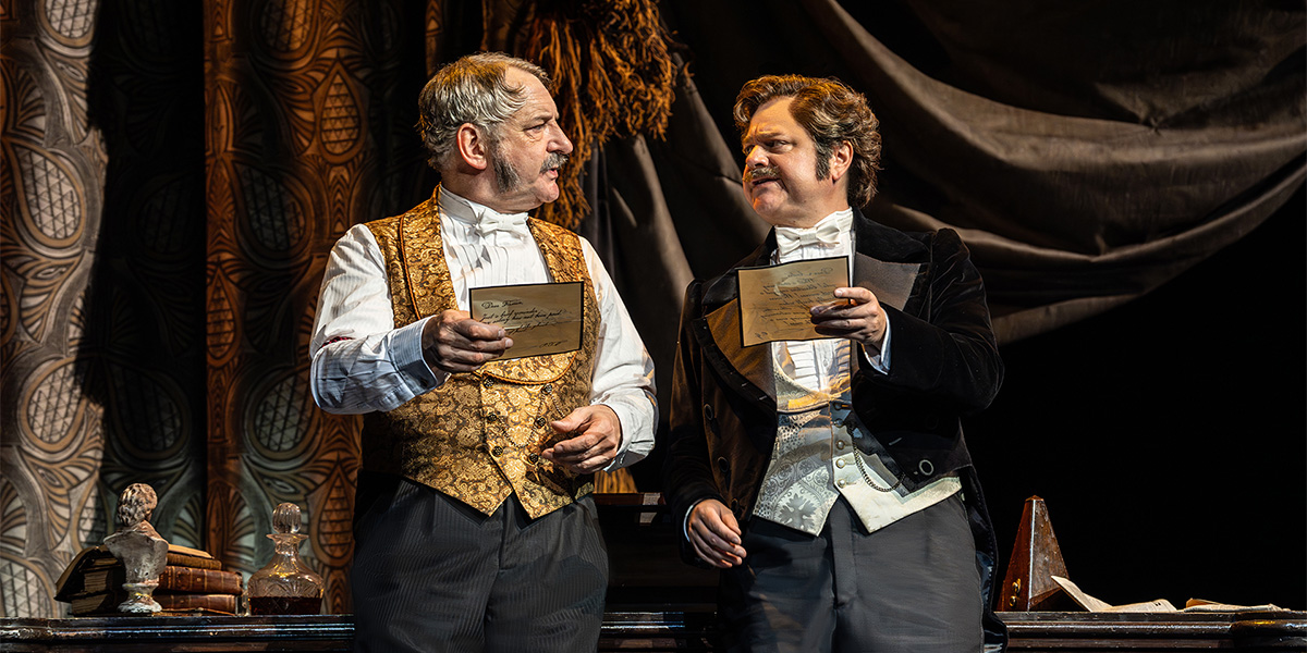 Two men in formal attire hold letters, engaged in conversation against a richly textured backdrop.