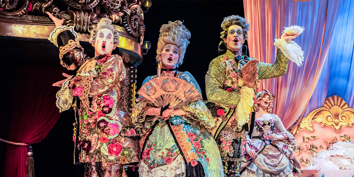Four performers in elaborate, floral costumes sing energetically, with vibrant colours and theatrical backdrops.