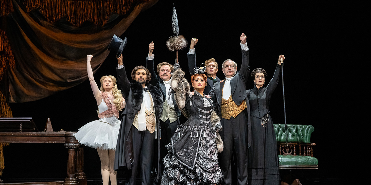 A group of six performers in theatrical costumes poses dramatically, raising their arms against a dark background.