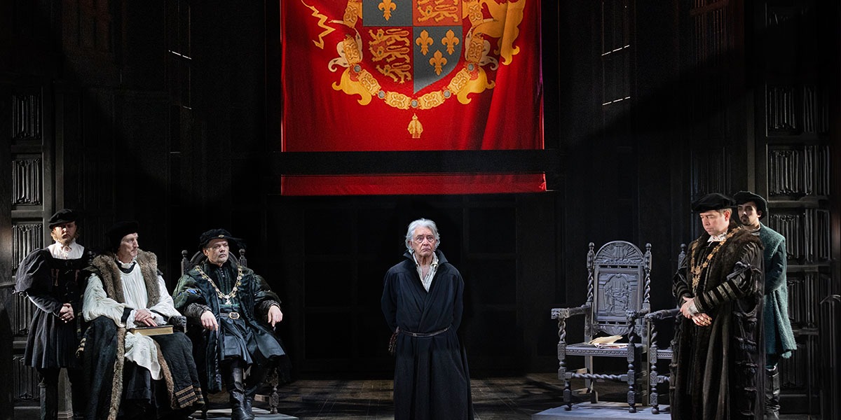 A stately room with a royal banner draped from the ceiling and stately chairs. To the left three men are sat looking at Martin Shaw, standing, in the centre
