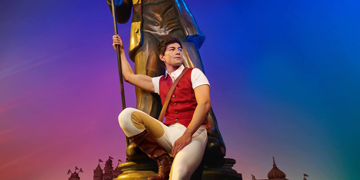 A performer in a green costume poses on stage, with a dark curtain backdrop.