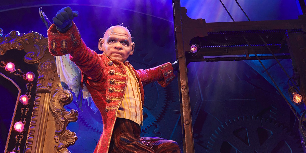 A green witch's hat with a textured surface, set against a dark, blurred theatre background.