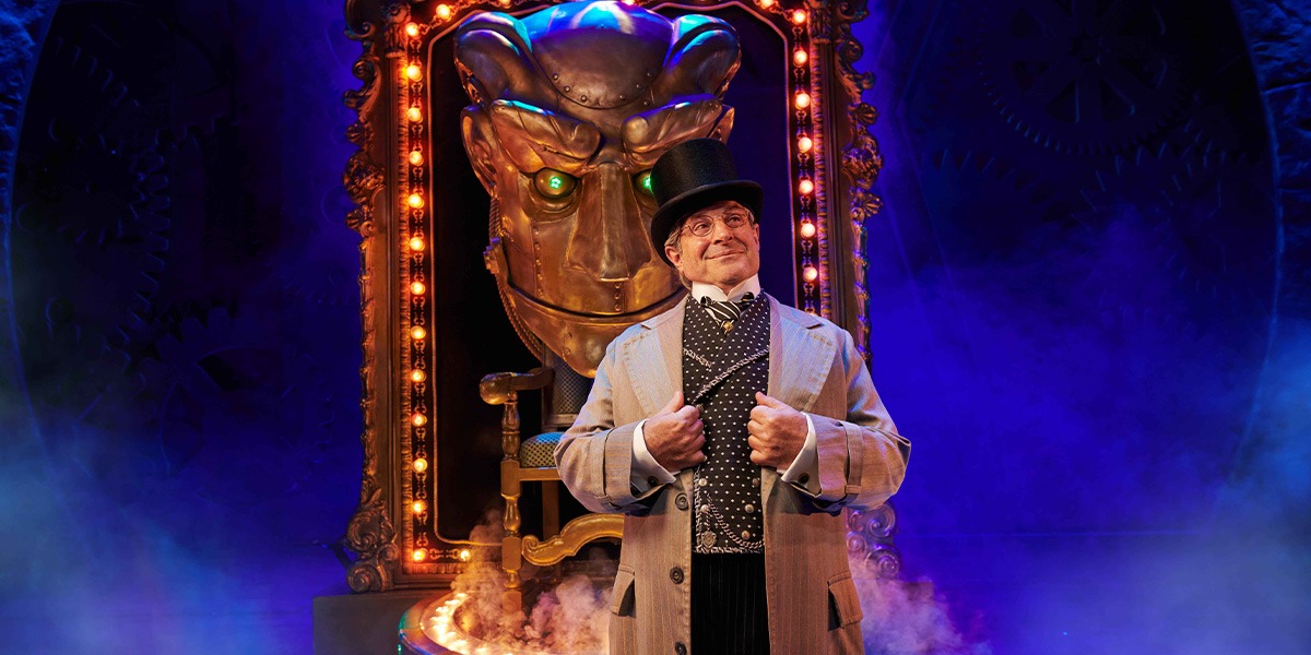 A performer in a green costume stands on stage, with dramatic lighting and a dark background.