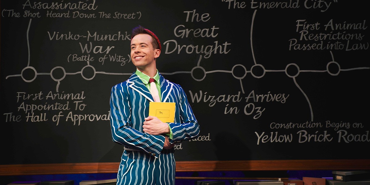 Ross Carpenter as Fiyero, wearing a green costume, stands on stage with dramatic lighting in the background.