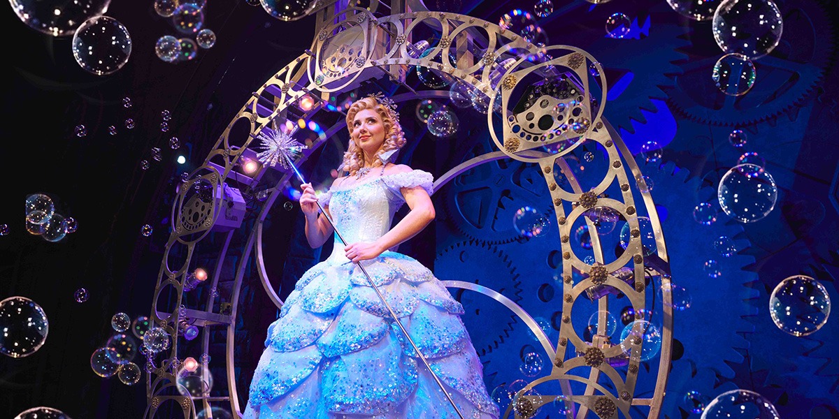 Zizi Strallen as Elphaba, wearing a green costume with a textured background of stage lights.