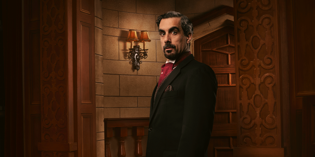 A man in a black suit and red tie stands confidently, with ornate wooden decor and warm lighting behind him.