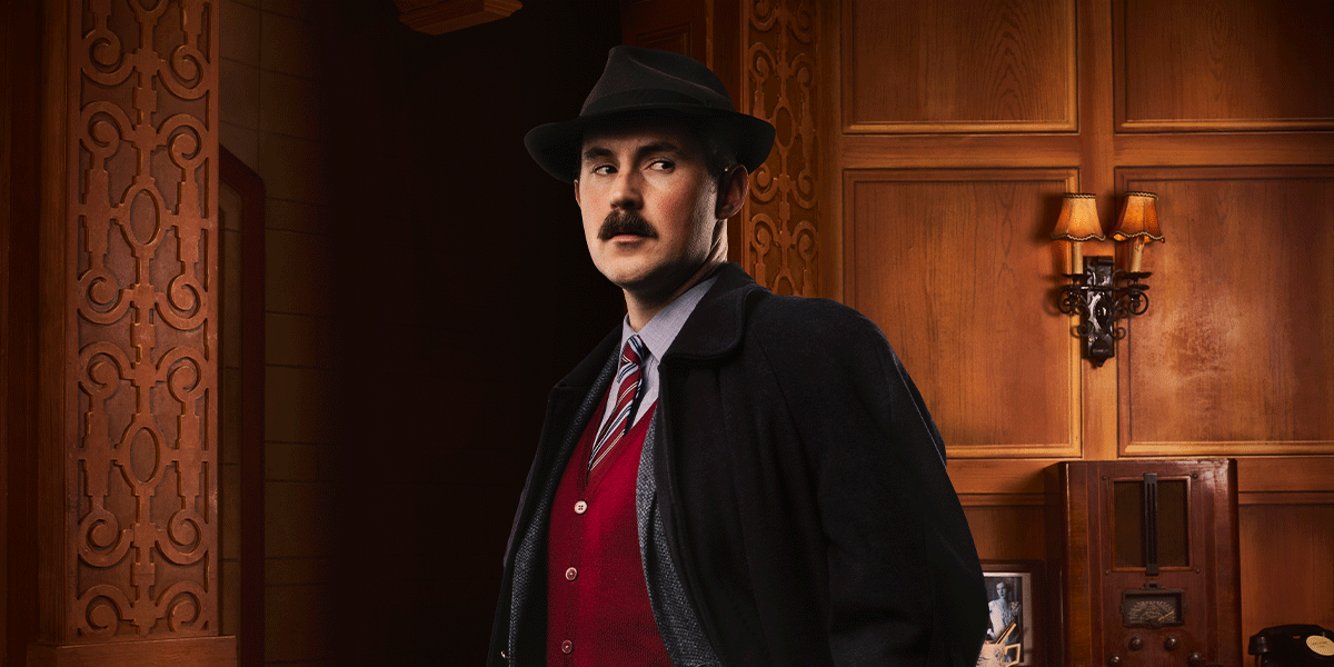 A man in a black hat and coat stands confidently, with a wooden panelled background and warm lighting.