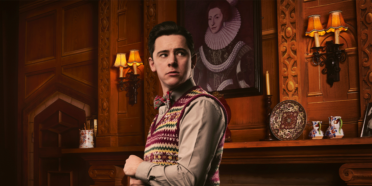 A young man in a patterned vest stands by a wooden mantel, looking thoughtfully over his shoulder.