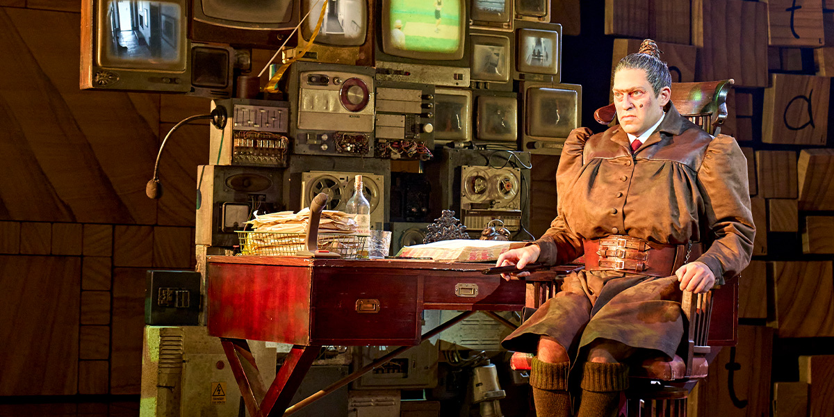 A stern character in a brown costume sits at a desk, surrounded by vintage televisions and paperwork.