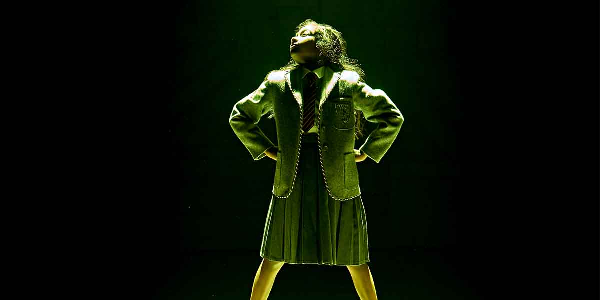 A person stands confidently in a green spotlight, wearing a grey school uniform.
