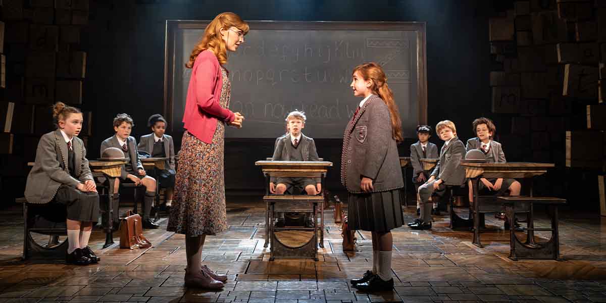 A child wearing a grey school uniform stands facing a teacher, surrounded by seated classmates in a classroom.