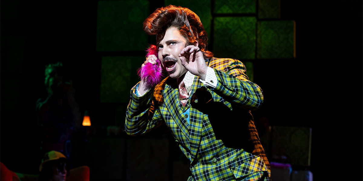 A man in a bright green and yellow checkered suit gestures animatedly while holding a pink object.