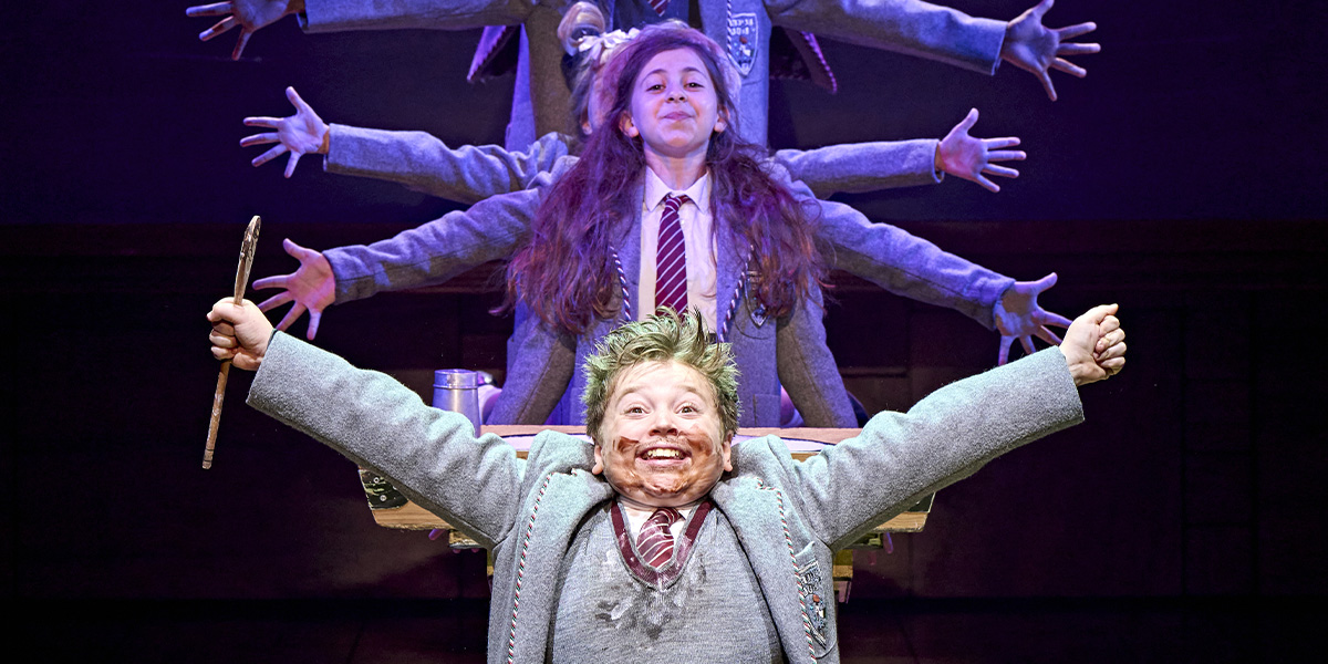 A child in a grey school uniform smiles widely, holding a stick, with multiple arms behind them.