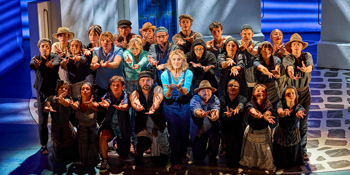A diverse group of performers in costumes, reaching out with hands, against a vibrant stage backdrop.
