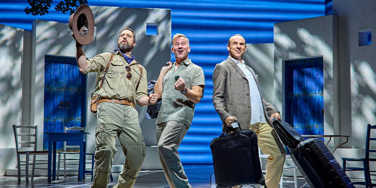 Three men stand on stage, smiling and posing with luggage, against a backdrop of blue and white.