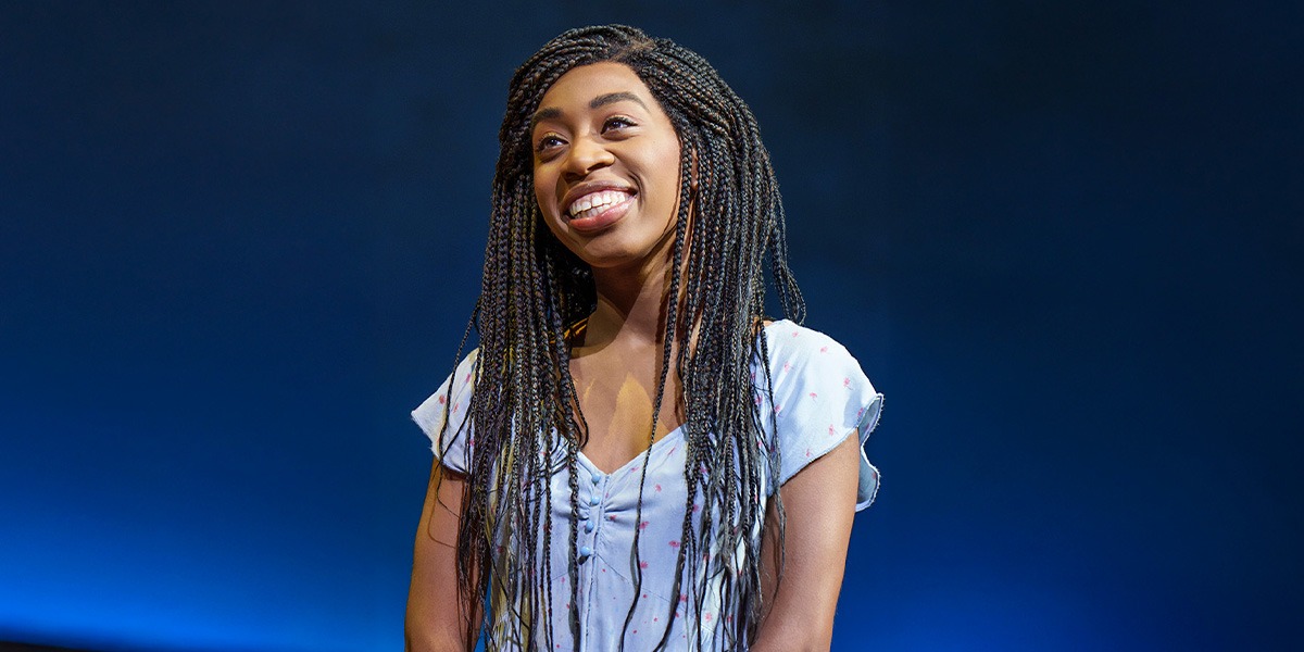 A woman with long braided hair smiles brightly, wearing a light blue top against a dark background.