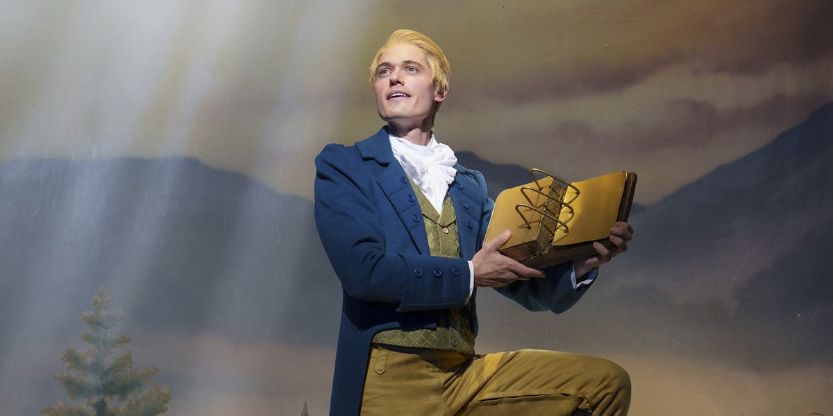 A man in a blue coat and yellow trousers holds a large book, posed against a scenic backdrop.
