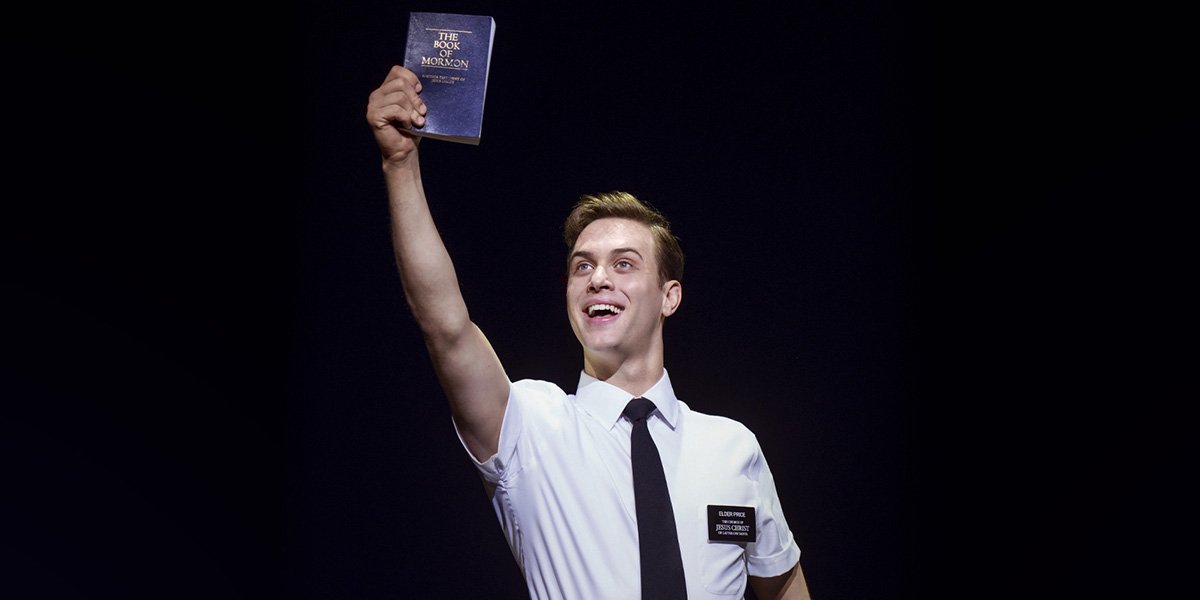 A young man in a white shirt and black tie holds up a book, smiling broadly against a dark background.