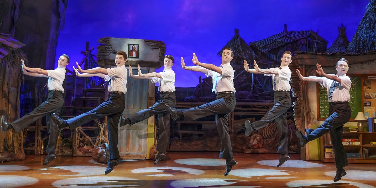 Five male performers in white shirts and black trousers dance energetically on stage against a colourful backdrop.