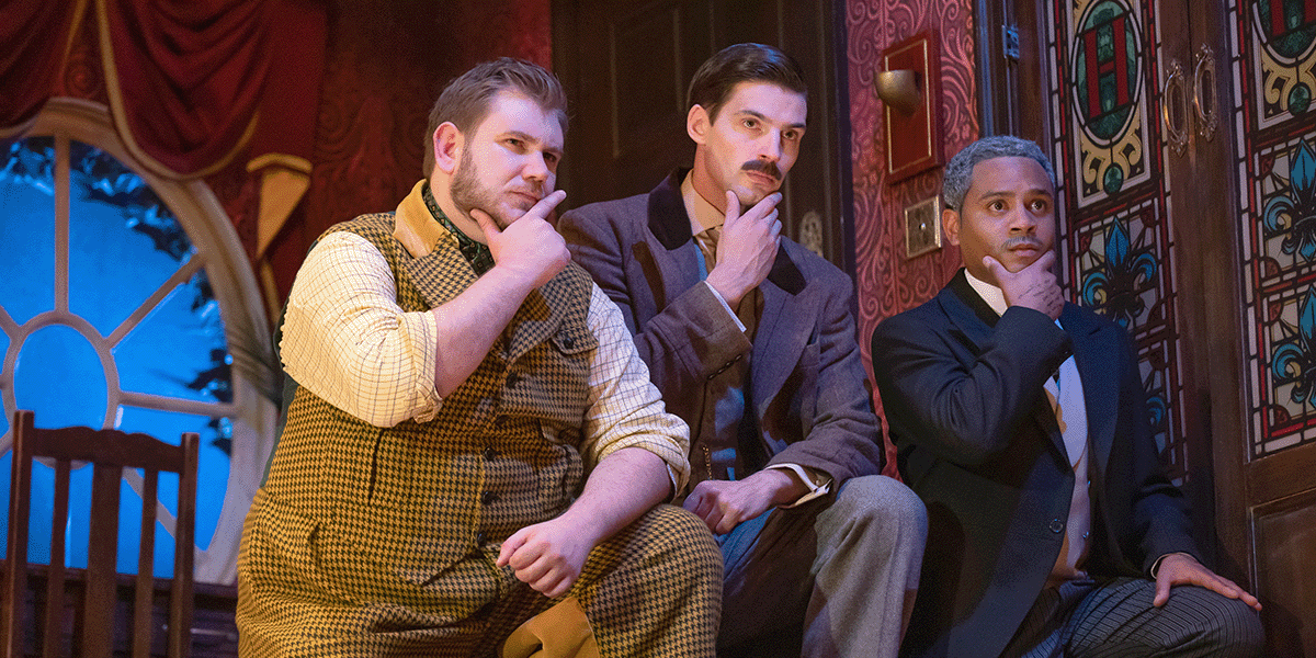 Three male actors in formal attire, expressing surprise, stand on a stage with theatrical props and curtains.
