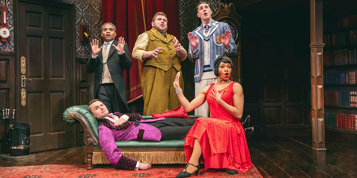 A group of actors in period costumes perform on stage, surrounded by theatrical props and a painted backdrop.