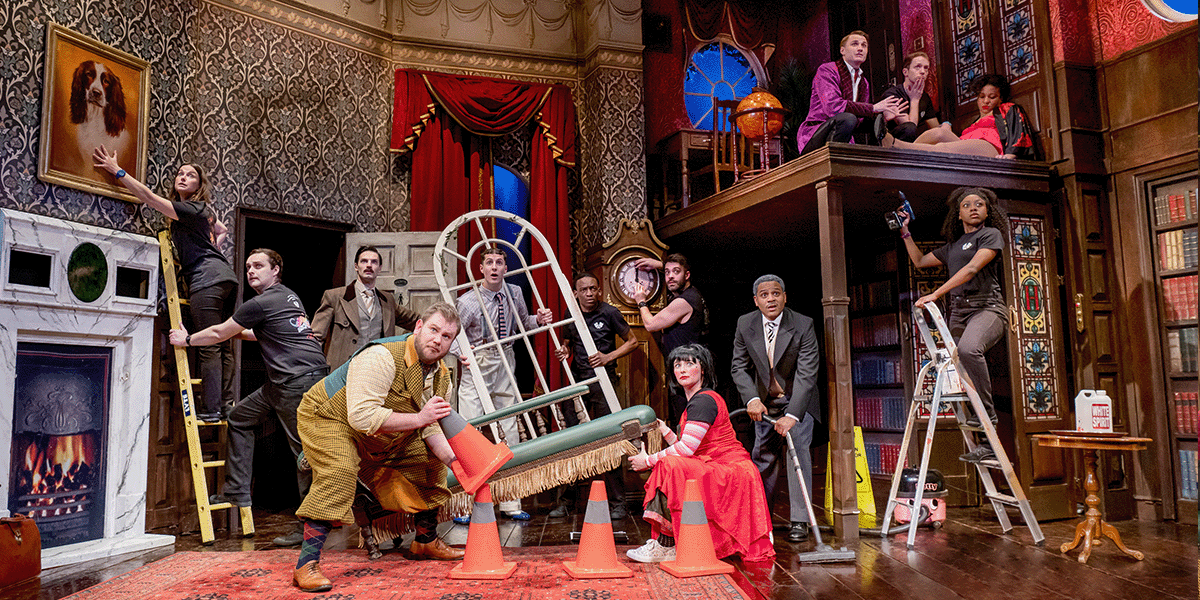 A group of actors in period costumes performs on a cluttered stage, with props scattered around.