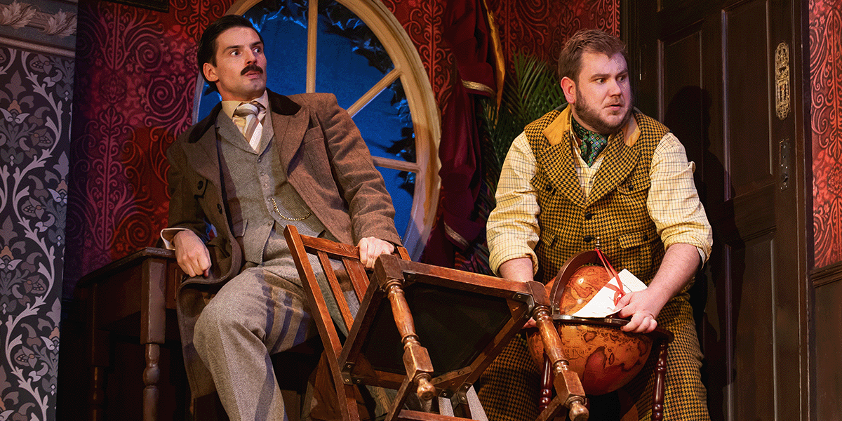 Two actors in formal attire, one in a tuxedo and the other in a suit, perform on stage.