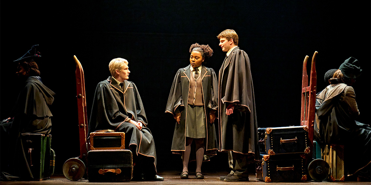 Five actors in costume pose together, with vibrant robes and accessories, against a dark theatrical backdrop.